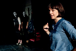Eine Frau steht im Fokus des Bildes. Sie wird von zwei Gestalten beobachtet, die eine Scream-Maske tragen.