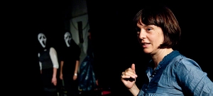 Eine Frau steht im Fokus des Bildes. Sie wird von zwei Gestalten beobachtet, die eine Scream-Maske tragen.