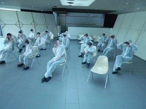Personen sitzen in Laborkitteln und mit schwarzen Taucherbrillen auf Stühlen in einem leeren Raum.