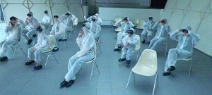 Personen sitzen in Laborkitteln und mit schwarzen Taucherbrillen auf Stühlen in einem leeren Raum.