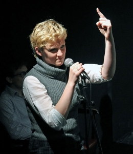 Eine Frau steht am Mikrofon. Sie zeigt mit dem linken Finger nach oben.