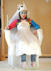 Ein verkleidetes Mädchen sitzt in einem weißen Sessel. Es hat die Augen blau geschminkt, trägt eine weiße Krone und ein weißes Kleid.