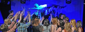 Eine junge Frau beim Crowdsurfing über einen Pulk von Jugendlichen. Der Raum ist blau beleuchtet.