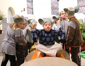 Ein Kind sitzt im Schneidersitz auf einem Tisch und schließt trotzig die Augen. Um das Kind herum stehen weitere Kinder. Alle tragen selbstgebastelte Mäuseohren auf dem Kopf.