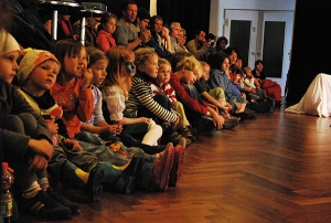 Kinder und Erwachsene sitzen in Reihen auf dem Fußboden und beobachten ein Theaterstück.
