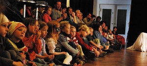 Kinder und Erwachsene sitzen in Reihen auf dem Fußboden und beobachten ein Theaterstück.