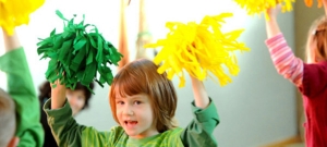 Ein Kind mit grüner Kleidung hält einen grünen und einen gelben Pompom in die Luft. Im Hintergrund stehen weitere Kinder mit Pompoms.