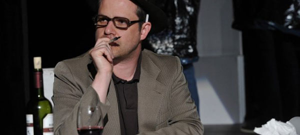 Ein Mann sitzt mit einem Glas Wein am Schreibtisch. Er hält einen Stift in der Hand und nimmt eine nachdenkliche Pose ein. Hinter seinem Ohr klemmt eine Zigarette.