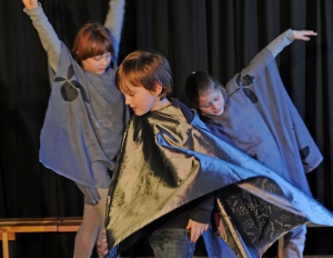Kinder tanzen auf einer Bühne. Sie tragen graue Umhänge.