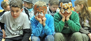 Eine Gruppe von Kindern sitzt in einem Klassenzimmer. Zwei der Kinder tragen selbstgebastelte Tiermasken.