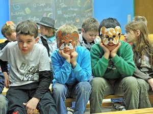 Eine Gruppe von Kindern sitzt in einem Klassenzimmer. Zwei der Kinder tragen selbstgebastelte Tiermasken.