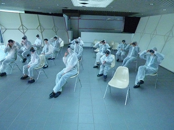 Menschen sitzen in Laborkitteln und mit Schutzbrillen auf Stühlen in einem leeren Raum.