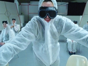 Eine Person in weißem Laborkittel und mit schwarzer Taucherbrille schaut in die Kamera. Im Hintergrund sitzen weitere Personen in Laborkitteln.
