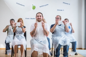 Mehrere Personen sitzen im Pulk auf Stühlen. Alle halten ein weißes Blatt Papier vor sich. Die Personen tragen weiße Schutzkittel.