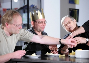 Drei Personen sitzen am Esstisch und schneiden Äpfel klein. Der Mann in der Mitte trägt eine selbstgebastelte, goldene Krone.