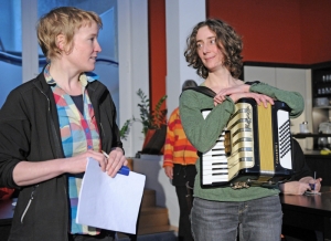 Zwei Theaterpädagoginnen vom TPZ stehen nebeneinander. Die linke Person hält Papiere in der Hand. die rechte Person trägt ein Akkordeon.