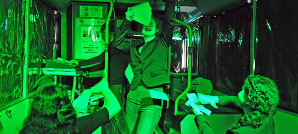 Eine Frau tanzt durch einen Bus. Sie hält mehrere Scheine in der Hand. Weitere Passagiere jubeln ihr zu. Der Bus ist grün beleuchtet, die Scheiben sind mit Folie verklebt.