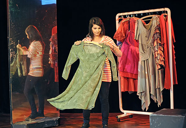 Eine Frau steht vor einem Spiegel und hält ein grünes Kleid in der Hand. Hinter ihr steht ein Kleiderständer, an dem mehrere Kostüme hängen.