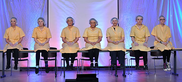 Viele Frauen sitzen nebeneinander auf einem langen Tisch. Sie tragen gelbe Kostüme und einen Haarschutz.