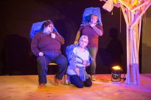 Drei Personen befinden sich auf einer Bühne. Sie tragen große, blaue Plastiktüten über dem Kopf.