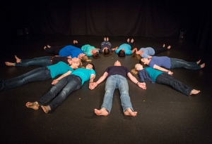 Menschen liegen mit dem Kopf nach innen im Kreis und fassen sich an den Händen. Sie tragen blaue T-Shirts.