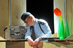 Eine Person in Polizeiuniform beugt sich über einen Holztresen und schläft. Neben ihr steht ein großes Radio und eine rote Tulpe aus Plastik.