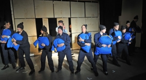 Viele Personen stehen nebeneinander auf einer Bühne. Sie sind gekleidet wie Häftlinge und halten große, blaue Säcke in den Händen. Hinter ihnen steht ein Polizist mit einer Pistole.