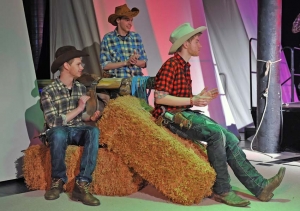 Drei junge Männer sitzen auf Strohballen. Die Männer sind als Cowboys verkleidet. Die Männer klatschen und schauen nach rechts aus dem Bild heraus.