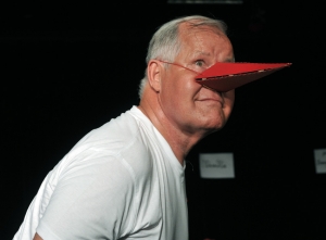 Ein Mann in weißem T-Shirt trägt einen roten Schnabel aus Papier auf der Nase. Der Hintergrund ist dunkel.