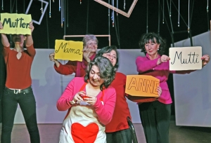 Fünf Frauen stehen auf einer Bühne. Die Frau vorne in der Mitte trägt eine Kochschürze mit einem großen roten Herz darauf. Die anderen Frauen stehen hinter ihr und halten Schilder in die Luft. Auf den Schildern steht jeweils ein Wort: "Mutter", "Mama", "Anne" und "Mutti".