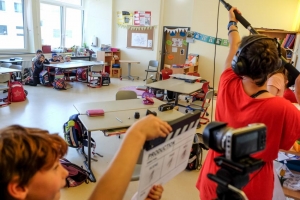 Kinder sitzen an einem Schultisch. Eine Kamera ist auf sie gerichtet. Im Vordergrund des Bildes steht eine Frau mit Kopfhörern und einer Tonangel in der Hand. Neben ihr steht ein Junge mit einer Filmklappe.