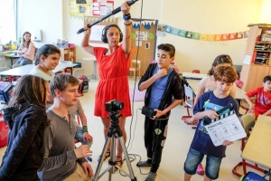 Das Foto wurde in einem Klassenzimmer aufgenommen. Eine Frau mit Kopfhörern hält eine Tonangel. Vor ihr steht eine Kamera. Kinder und ein Mann sitzen und stehen im Kreis um die Kamera herum. Ein Kind hält eine Filmklappe in der Hand.