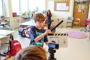 Das Foto wurde in einem Klassenzimmer aufgenommen. Ein Junge hält eine Filmklappe vor eine Kamera. Im Hintergrund stehen und sitzen drei weitere Schulkinder und schauen zu.