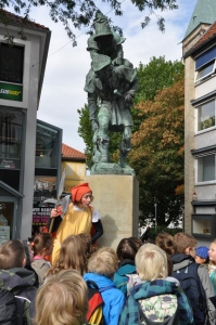 Eine Frau im Kostüm gibt eine Stadtführung für Kinder. Sie stehen vor dem Huckup in Hildesheim.