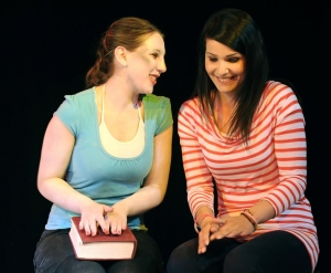 Zwei junge Frauen sitzen nebeneinander und unterhalten sich lachend. Die linke Frau hat ein großes, rotes Buch auf ihrem Schoß liegen.