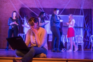 Eine junge Frau sitzt am Rand einer Bühne und schaut konzentriert auf einen Laptop. Im Hintergrund laufen Frauen mit Smartphones umher.