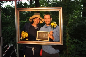 Zwei Männer in Verkleidung schauen durch einen goldenen Bilderrahmen. Hinter ihnen befinden sich Bäume. Die Männer halten eine kleine Tafel hoch. Darauf wurde mit Kreide geschrieben: "16/.12"