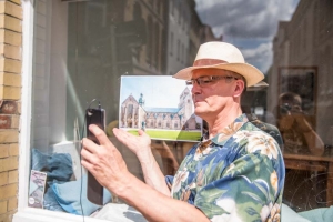 Ein Mann steht vor einem Schaufenster und filmt sich mit dem Handy. Er zeigt auf ein Foto, das hinter ihm am Schaufenster klebt. Auf dem Foto ist der Hildesheimer Dom abgebildet. Im Schaufenster spiegelt sich eine Person, die den Mann fotografiert.