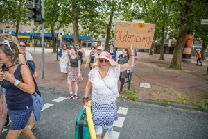 Viele Personen laufen vom Hindenburgplatz in Hildesheim aus über die Straße. Eine Frau mit Sonnenhut, Poolnudel und einem Koffer in der Hand hält ein Pappschild in der Hand. Auf dem Schild steht: "Oldenburg".