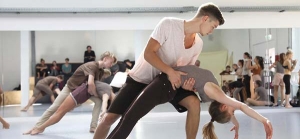 Junge Menschen tanzen paarweise in einem Tanzsaal. Die Männer halten die Frauen fest, die sich nach hinten lehnen. Im Hintergrund stehen viele Menschen und schauen zu.