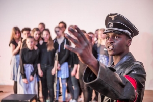 Eine farbige Person in SS-Uniform deutet mit der Hand nach vorne. Im Hintergrund steht ein Pulk von jungen Menschen und schaut die Person an.