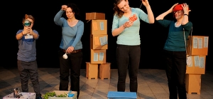 Vier Personen stehen auf einer Bühne und haben gefüllte Kisten vor sich stehen. Sie betrachten die Dinge aus den Kisten: Einen Pokal, eine kleine Tasse, einen Hut. Im Hintergrund sind Kartons gestapelt. Auf den Kartons sind Zettel angebracht.