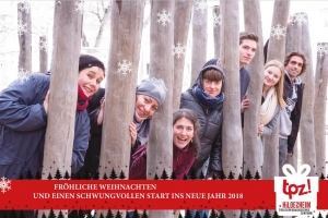 Die Mitarbeitenden des TPZ schauen an Baumstämmen vorbei in die Kamera. Unter dem Foto befindet sich ein roter Banner. Darauf steht: "Fröhliche Weihnachten und einen schwungvollen Start ins neue Jahr 2018". Rechts daneben befindet sich das Logo vom TPZ in einem weißen Geschenk.