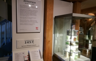 Eine Vitrine in einem Museum. In der Vitrine sind leuchtende Becher aufeinander gestapelt. Neben der Vitrine befindet sich ein eingerahmtes Schild mit einem Text. Über dem Text steht: "Zeitkapseln aus der freien Kultur". Unter dem Schild ist ein Zähler angebracht. Der Zähler zeigt die Zahl 3457. Darunter steht: "Tage bis zur Öffnung der Zeitkapseln".