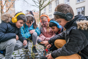 Fünf Kinder hocken auf einer Straße mit Kopfsteinpflaster. Die Kinder tragen Kopfhörer in unterschiedlichen Farben. Die Kabel der Kopfhörer laufen in der Mitte zusammen. Alle Kinder schauen auf die Kabel.