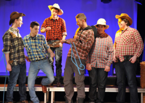 Das Foto entstand während einer Theaterraufführung. Jugendliche stehen auf einer Bühne. Sie tragen Hemden mit Karomuster und Cowboyhüte. Der Mann in der Mitte hält ein blaues Seil.
