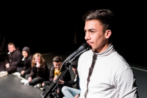 Ein junger Mann steht an einem Mikrofon. Links neben ihm sitzen mehrere Jugendliche auf dem Boden.