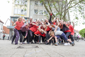 Viele Personen posieren auf einem Platz in Hildesheim. Die Personen tragen Partyhütchen, rote T-Shirts und schwarze Hosen. Die Personen lachen und strecken ihre Arme aus.