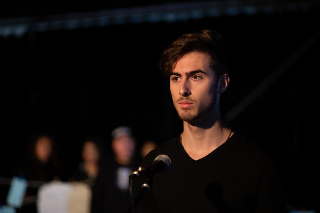 Ein junger Mann steht vor einem Mikrofon. Der Hintergrund ist dunkel.