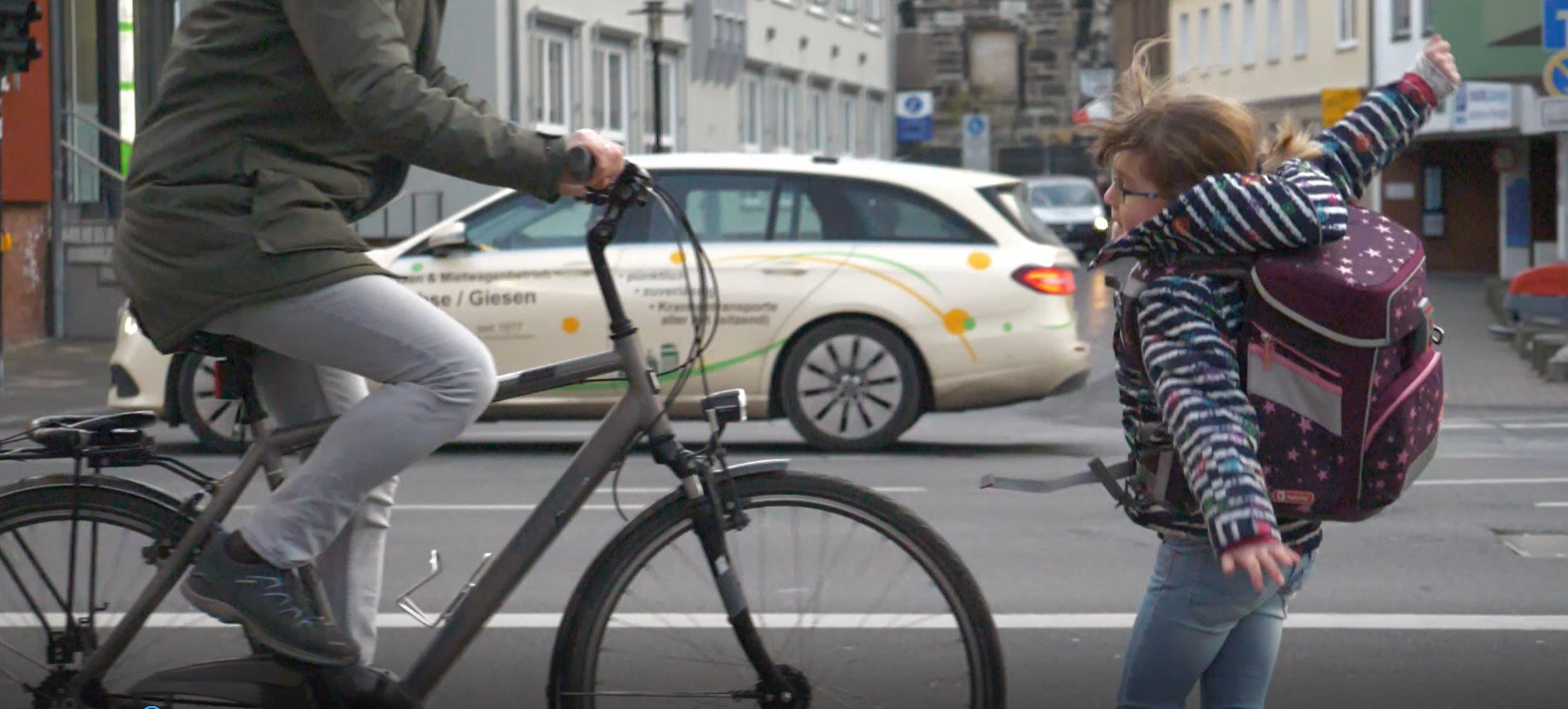 Ein Fahrradfahrer fährt über eine Straße. Ein Mädchen mit Schultasche weicht dem Fahrrad aus.